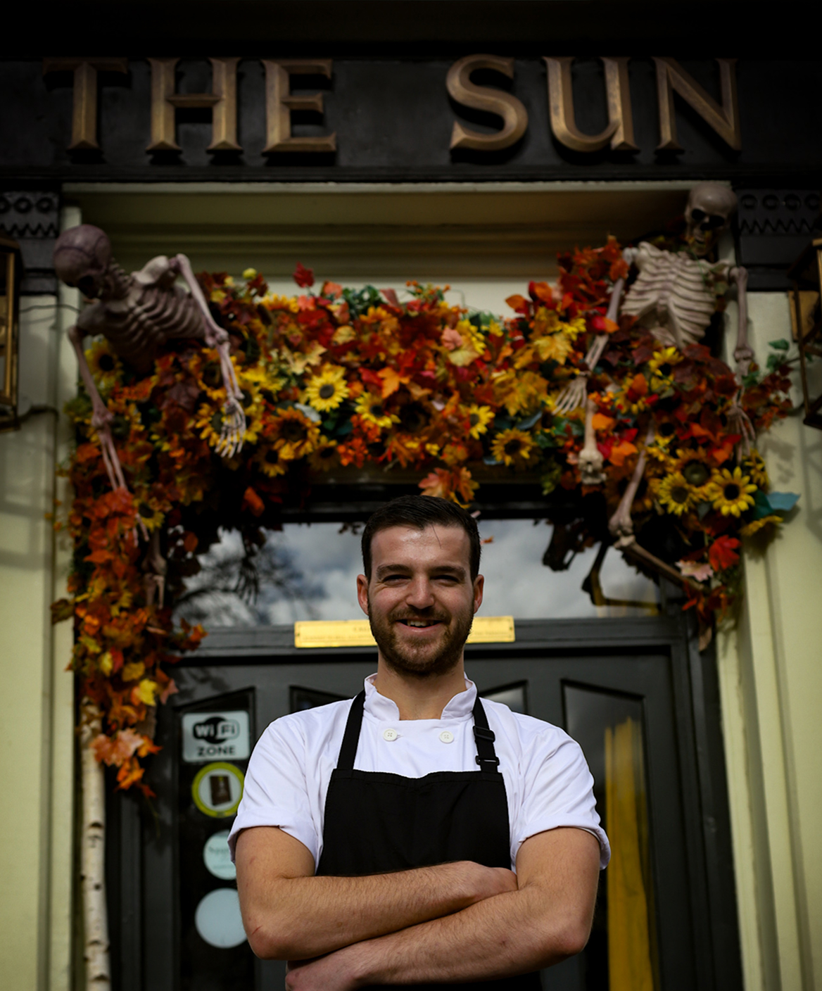 An image of a man in chefs uniform standing in front of a building with the words the sun above the door